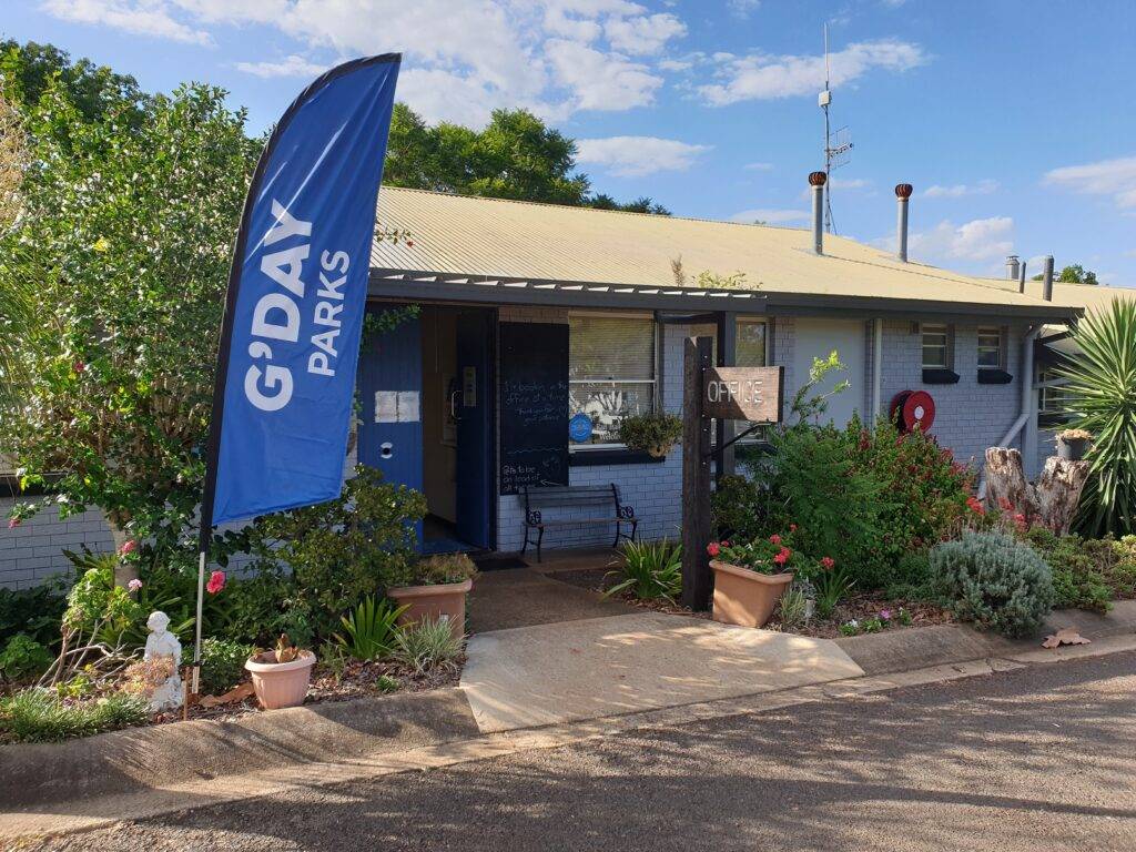 external view of caravan park reception area with G'day Parks flag and open reception door with planter boxes surrounding